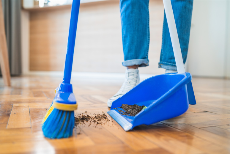 A person standing behinda broom and dustpan sweeping up dirt. 